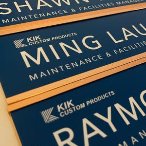 three blue nameplates with white text, each displaying the name "SHAWN" and the company name "CUSTOM PRODUCTS" along with the tagline "MAINTENANCE & FACILITIES MANAGER".