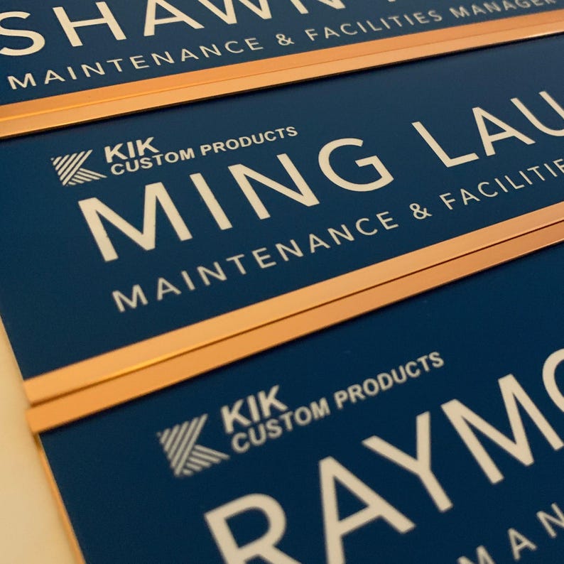 three blue nameplates with white text, each displaying the name "SHAWN" and the company name "CUSTOM PRODUCTS" along with the tagline "MAINTENANCE & FACILITIES MANAGER".