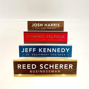 May include: Four wooden desk nameplates with gold trim. The top nameplate reads "Josh Harris - Evil Dies Tonight -" in silver lettering. The second nameplate reads "Dominic Pelpola - Supervisor / Sith Lord -" in silver lettering. The third nameplate reads "Jeff Kennedy - Sr. Equipment Engineer -" in silver lettering. The bottom nameplate reads "Reed Scherer - Businessman" in white lettering.