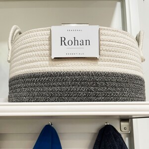 Puede incluir: Una cesta de almacenamiento de cuerda blanca y gris con una etiqueta plateada que dice "SEASONAL Rohan ESSENTIALS".