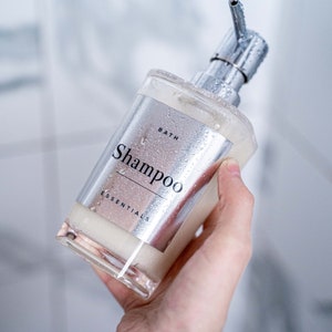 Puede incluir: Una botella de bomba plateada con una etiqueta transparente que dice "Bath Essentials Shampoo". La botella est&aacute; mojada y tiene gotas de agua.