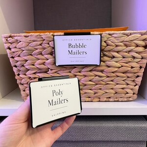 Puede incluir: Dos etiquetas blancas con texto negro y bordes negros, unidas a una cesta de mimbre. La etiqueta superior dice "Office Essentials Bubble Mailers Shipping". La etiqueta inferior dice "Office Essentials Poly Mailers Shipping".