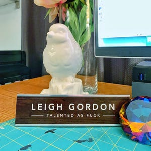 a wooden nameplate with the name 'Leigh Gordon' on it, placed on a table with a vase of flowers and a computer monitor in the background.