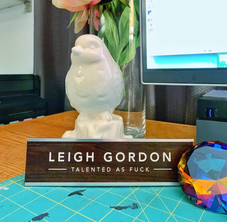 a wooden nameplate with the name 'Leigh Gordon' on it, placed on a table with a vase of flowers and a computer monitor in the background.