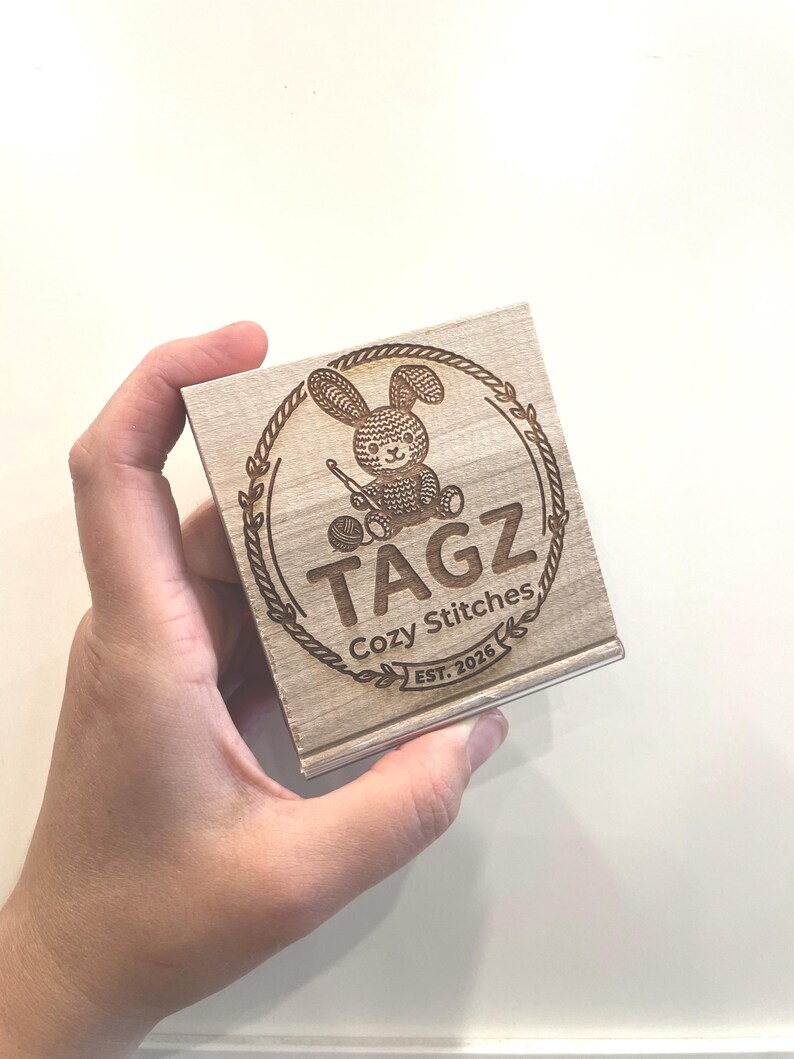 May include: A square wooden box featuring a laser-engraved design of a rabbit holding a crochet hook next to a ball of yarn. The text reads TAGZ Cozy Stitches EST. 2026, all enclosed within a circular laurel wreath border on the light wood surface.