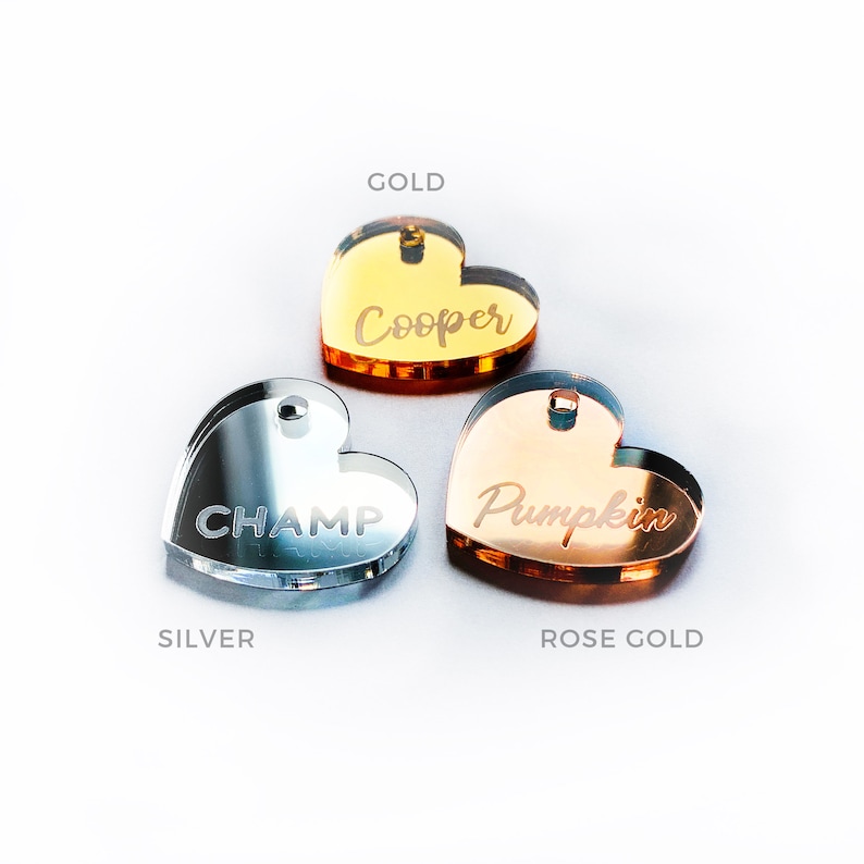 May include: Three heart-shaped pet tags in gold, silver, and rose gold. The gold tag says "Cooper", the silver tag says "Champ", and the rose gold tag says "Pumpkin". Each tag has a small hole for attaching to a collar.
