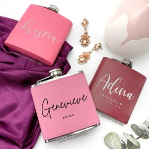 Könnte beinhalten: Drei personalisierte Flachmänner in Rosa und Bordeaux, mit silbernen Akzenten. Einer ist mit "Kristin" graviert, ein anderer mit "Adina, Bridesmaid 20 08 22", und der dritte mit "Genevieve, Bride". Goldfarbene Ohrringe sind ebenfalls zu sehen.