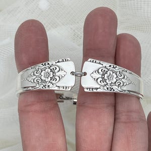 May include: A silver-toned bracelet crafted from two segments of ornate metal cutlery handles. Each segment features a detailed embossed floral pattern with scrolling leaves. The two pieces are connected by a small circular metal jump ring.
