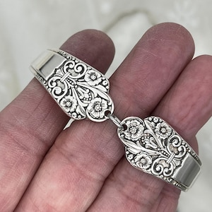 May include: A silver-toned bracelet crafted from two repurposed spoon handles. Each handle features an intricate embossed floral pattern with detailed petals and leaves, connected by a small metal jump ring to form a continuous link design.