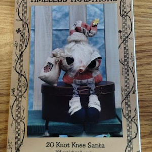 May include: A Timeless Traditions 20 Knot Knee Santa pattern booklet featuring a 30.48 cm weighted Santa doll with a red and white checkered hat, a white beard, a small sack, and dangling legs with black shoes and white socks, framed by a vine border.