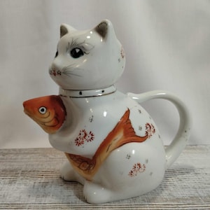 May include: A white ceramic cat-shaped teapot, clutching an orange fish. The teapot features a handle and spout, adorned with red floral patterns and a gold-coloured collar. A charming decorative item.