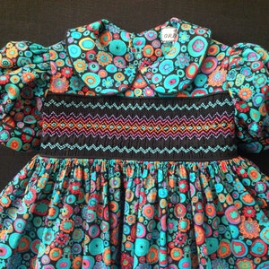 May include: A colorful baby dress with a floral pattern in teal, orange, and pink on a black background. The dress features a Peter Pan collar, puffed sleeves, and a smocked bodice with a geometric design.