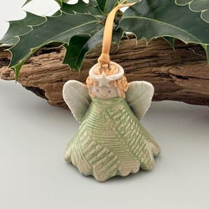 May include: A ceramic angel ornament featuring a light green dress with a textured fern leaf pattern, white wings, and a small star halo. The figure is suspended by a thin orange ribbon and is positioned in front of dark green holly leaves.