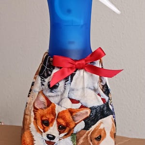 May include: A blue spray bottle with a white nozzle is wrapped in a fabric cover featuring various dog breeds. A red satin bow adorns the top. The fabric showcases corgis, beagles, and other dogs.