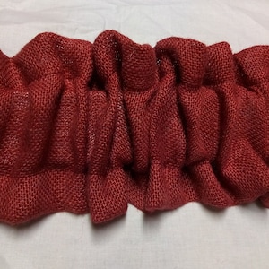 May include: Close-up of a ruffled, burgundy-colored burlap fabric. The fabric is tightly woven and has a textured appearance. The ruffled design adds a decorative element, suitable for home decor or craft projects.