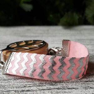 May include: Pink bracelet with a silver chevron pattern. The bracelet has a silver clasp and end caps. The background is a blurred view of a wooden surface and greenery.