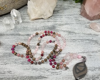 May include: A pink, brown, and red beaded necklace with a silver pendant. The necklace is laid out on a white wooden surface with pink and white crystals.