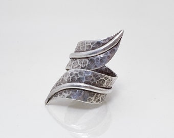 May include: A silver ring featuring a spiraling, wrap-around design. The metal surface displays a dark, oxidized, hammered texture with a network of fine cracks, contrasted by smooth, polished silver edges that define the leaf-like shape.