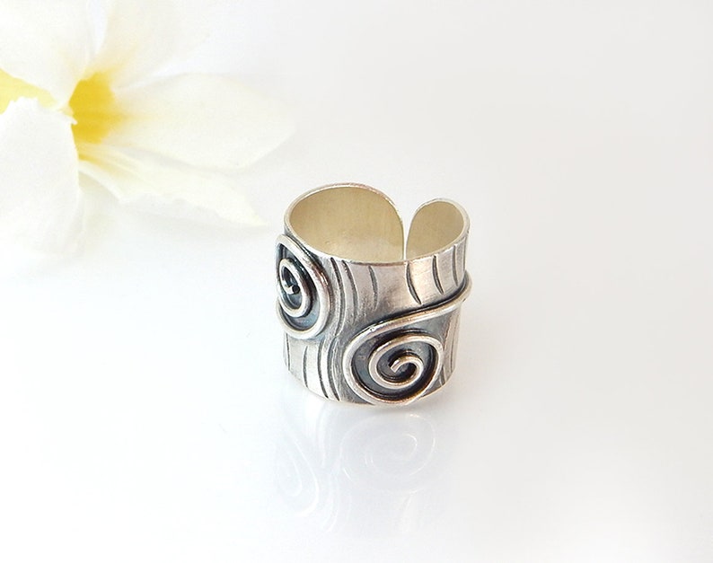Op de afbeelding: Een zilveren ring met een getextureerd oppervlak en twee spiraalontwerpen. De ring is breed en heeft een licht afgeronde vorm.