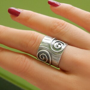 Op de afbeelding: Een zilveren ring met een brede band met een getextureerd ontwerp en twee spiraalpatronen.