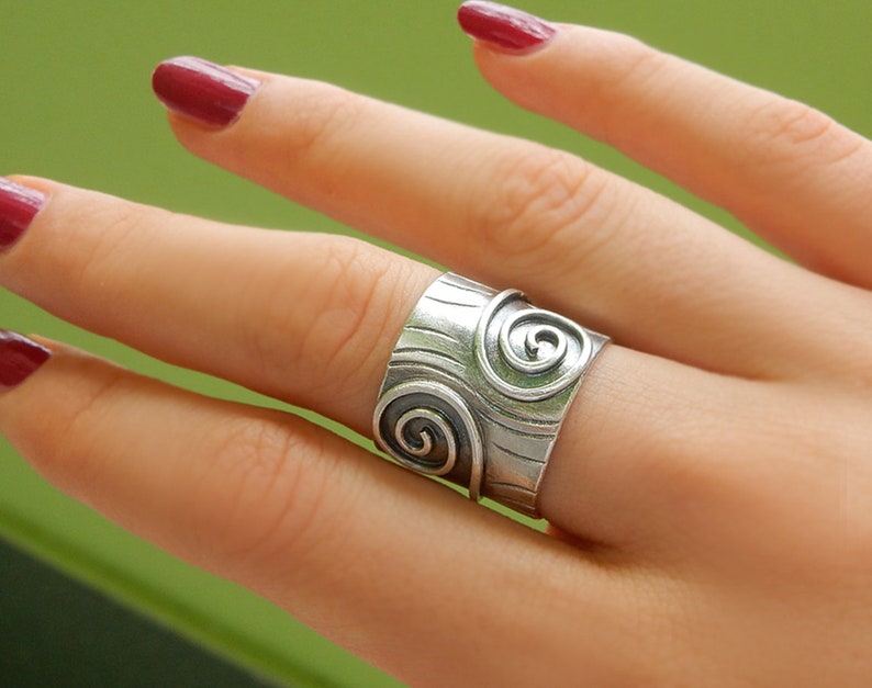 Op de afbeelding: Een zilveren ring met een brede band met een getextureerd ontwerp en twee spiraalpatronen.