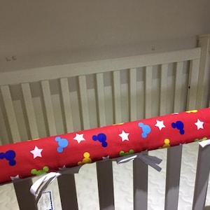 May include: A white crib with a red and white crib bumper featuring a Mickey Mouse pattern. The bumper has blue, yellow, and green accents.