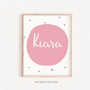 May include: A pink and white printable art print with the name "Kiara" in a white cursive font on a pink circle surrounded by gray stars. The print has a wooden frame.