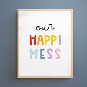 May include: A framed print with the words "our HAPPY MESS" in colourful, playful lettering. The text is set against a white background, framed in a light wood frame, and mounted on a blue wall.