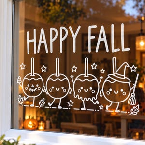 Op de afbeelding: Een witte raamsticker met de tekst HAPPY FALL boven vier cartoon-kandijappels met gezichtjes. Het ontwerp bevat sterren en bladeren, waarbij één appel een heksenhoed draagt en een bezem vasthoudt, alles in eenvoudige lijntekeningen.