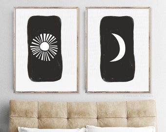 May include: Two black and white minimalist art prints with a sun and moon design. The sun print has a white sun with rays radiating out from it. The moon print has a white crescent moon. Both prints are framed in a light wood frame.