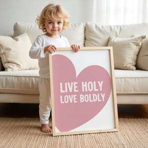 May include: A framed art print with a light wood frame. The print features a large pink heart on a white background with the words "LIVE HOLY LOVE BOLDLY" in white text. The art is held by a child.