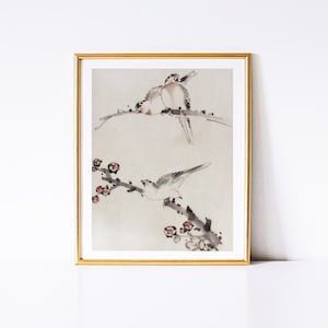 May include: A framed watercolor painting of two birds perched on a branch with delicate pink blossoms. The birds are painted in shades of gray and white, with subtle details of their feathers. The background is a soft, muted gray.