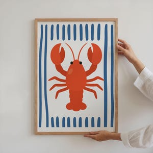 Puede incluir: Un cuadro enmarcado con una ilustraci&oacute;n de una langosta roja sobre un fondo blanco con rayas azules. La obra de arte est&aacute; enmarcada en un marco de madera marr&oacute;n claro, evocando un tema costero o n&aacute;utico.