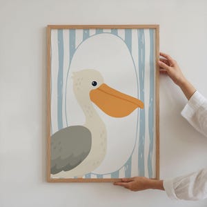 Puede incluir: Una impresi&oacute;n art&iacute;stica enmarcada con un pel&iacute;cano de dibujos animados. El pel&iacute;cano tiene un gran pico naranja y un ala gris, sobre un fondo de rayas verticales azules y blancas dentro de una forma ovalada. El marco es de madera clara.