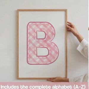 May include: A framed wall art print featuring a large, capital letter B in a pink and white gingham pattern. The letter has a white dashed border resembling stitching. The frame is light-colored wood. The text below states: Includes the complete alphabet (A-Z).
