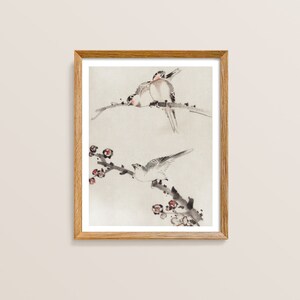 May include: A framed watercolor painting of two birds perched on a branch with delicate pink blossoms. The birds are painted in shades of gray and white, with subtle hints of pink and brown. The background is a soft, light gray.