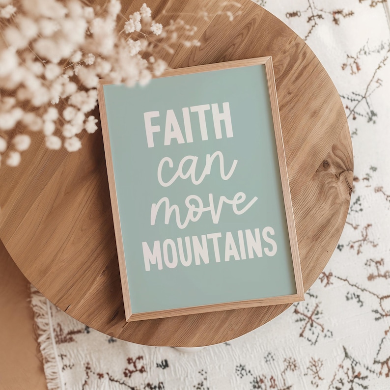 May include: A framed print with the words "FAITH can move MOUNTAINS" in white script on a light teal background. The frame is light brown. The print is on a wooden table with a white patterned rug.