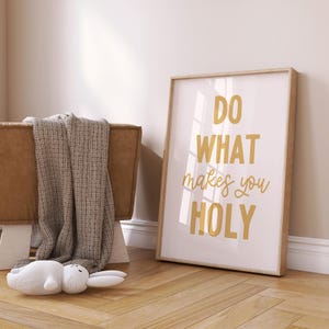 May include: A framed print with the text "DO WHAT makes you HOLY" in gold lettering. The print is leaning against a wall on a wooden floor. A brown bench with a gray blanket and a white stuffed bunny are in the foreground.