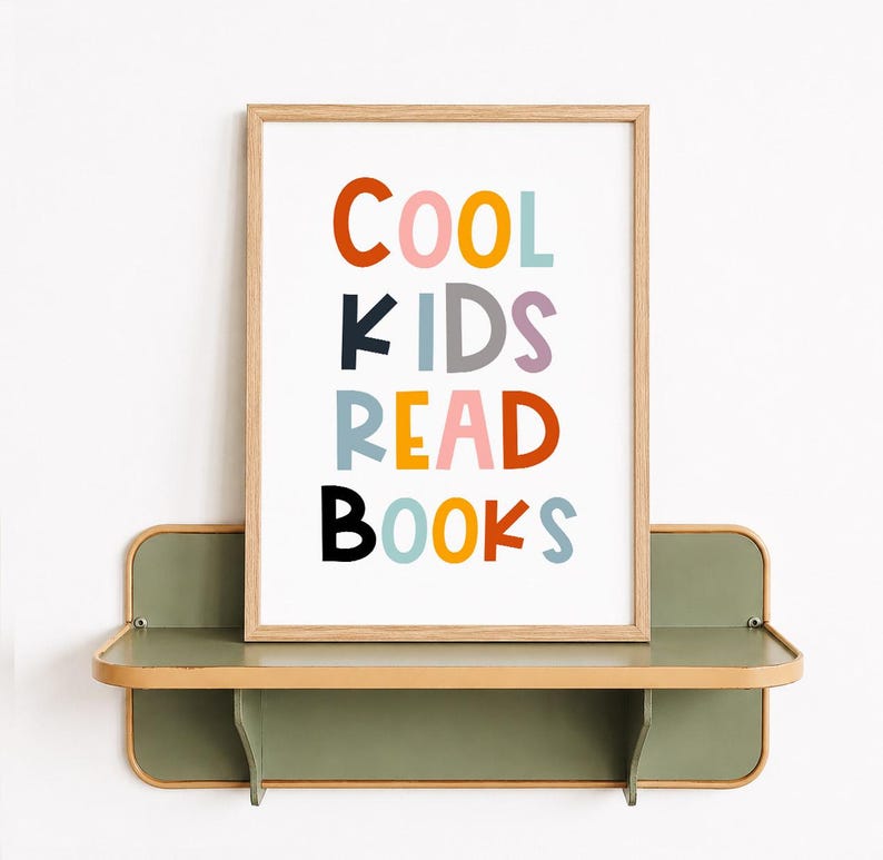 May include: A rectangular wooden frame containing a white print with the multicolored text COOL KIDS READ BOOKS. The letters are displayed in various colors including orange, pink, yellow, blue, grey, purple, and black, set against a white background.