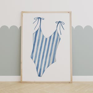May include: Watercolor illustration of a blue and white striped swimsuit. The one-piece swimsuit has a V-neck and tie straps. The artwork is framed in a light wood frame and displayed on a wooden floor.