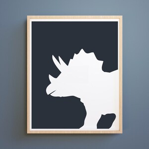 May include: A white silhouette of a triceratops dinosaur on a dark blue background. The image is framed in a light wood frame.