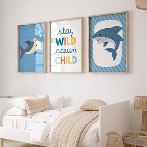 Puede incluir: Tres l&aacute;minas enmarcadas con tem&aacute;tica n&aacute;utica. Las l&aacute;minas presentan tiburones de dibujos animados y el texto "Stay Wild Ocean Child" en letras coloridas. Los marcos son de madera clara con fondos azules y blancos. La obra de arte se exhibe sobre una cama blanca.