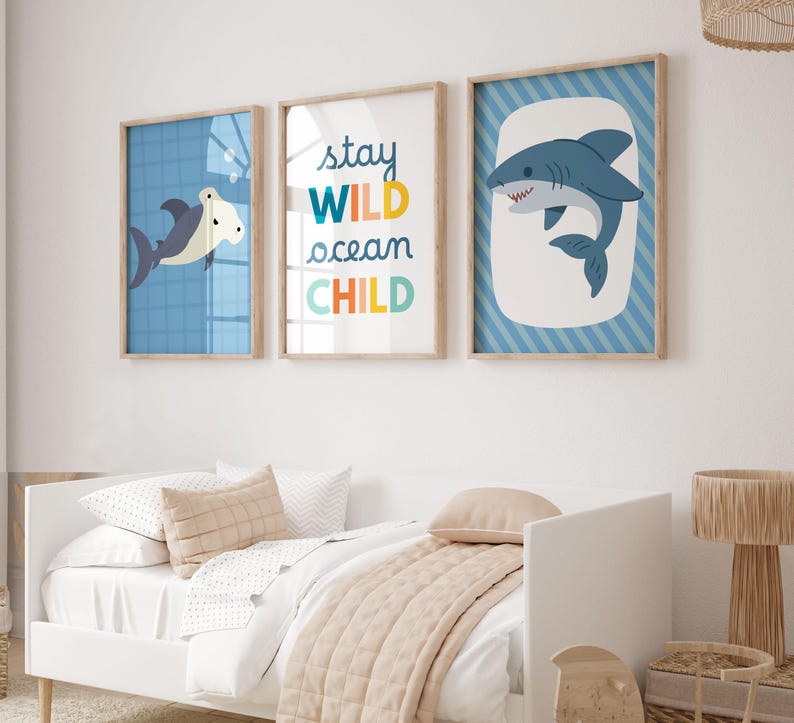 Puede incluir: Tres l&aacute;minas enmarcadas con tem&aacute;tica n&aacute;utica. Las l&aacute;minas presentan tiburones de dibujos animados y el texto "Stay Wild Ocean Child" en letras coloridas. Los marcos son de madera clara con fondos azules y blancos. La obra de arte se exhibe sobre una cama blanca.