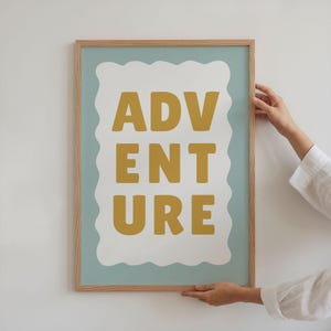 Puede incluir: Un cuadro enmarcado con la palabra "ADVENTURE" en letras de bloque amarillo mostaza. El texto est&aacute; sobre una forma blanca festoneada contra un fondo azul claro, todo dentro de un marco de madera natural.