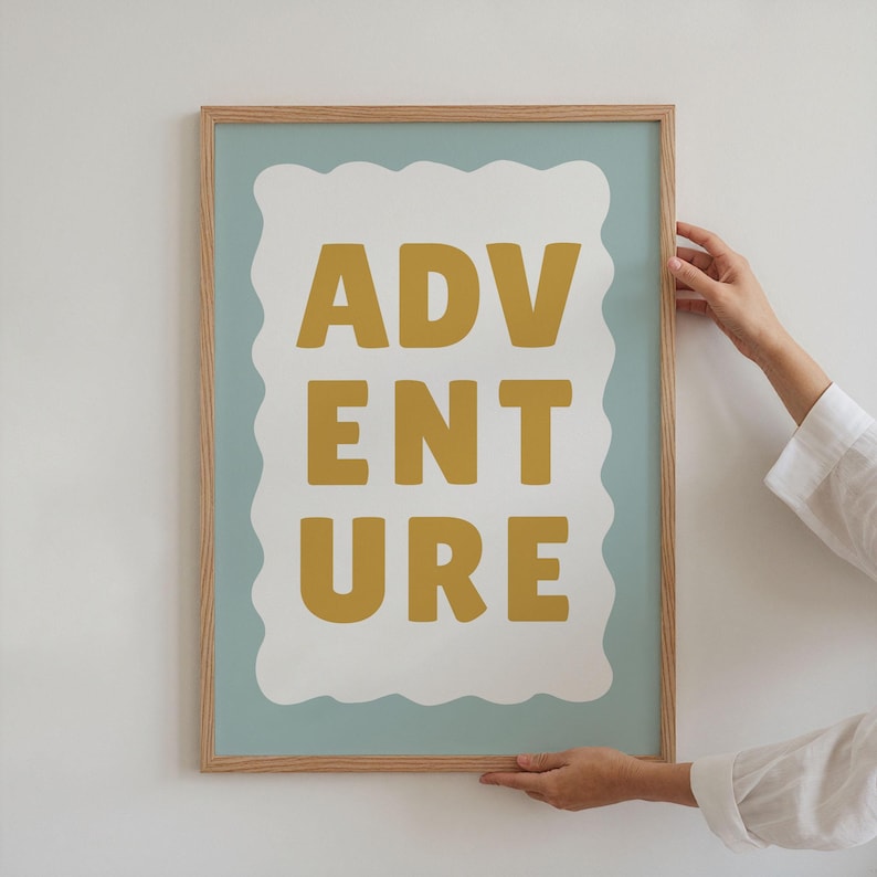 Puede incluir: Un cuadro enmarcado con la palabra "ADVENTURE" en letras de bloque amarillo mostaza. El texto est&aacute; sobre una forma blanca festoneada contra un fondo azul claro, todo dentro de un marco de madera natural.