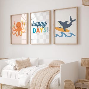 Puede incluir: Tres impresiones art&iacute;sticas enmarcadas con tem&aacute;tica de playa. Una impresi&oacute;n presenta un pulpo naranja sosteniendo conos de helado, otra dice "happy DAYS!", y la &uacute;ltima muestra un tibur&oacute;n surfeando sobre una tabla amarilla sobre olas azules. Las impresiones est&aacute;n en un marco de madera clara.