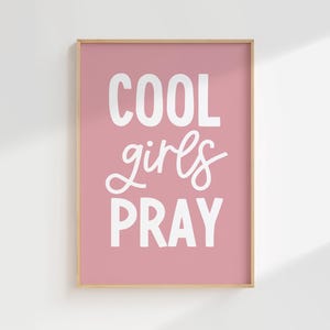 May include: A framed print with the words "COOL girls PRAY" in white lettering on a pink background. The frame is a light brown color. The print is displayed on a white wall.