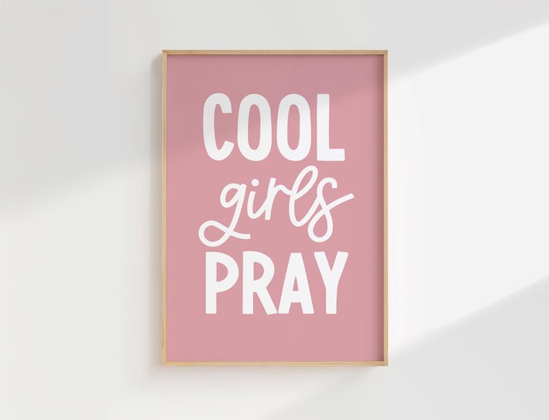 May include: A framed print with the words "COOL girls PRAY" in white lettering on a pink background. The frame is a light brown color. The print is displayed on a white wall.
