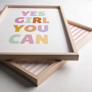 May include: Two light-colored wooden frames containing prints. The top print features the text YES GIRL YOU CAN in pastel colors including green, pink, orange, purple, and yellow. The bottom print shows a pattern of vertical pink and white stripes.
