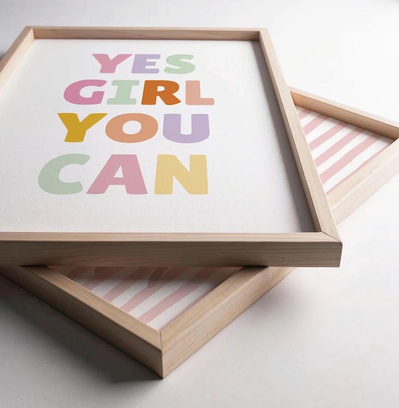 May include: Two light-colored wooden frames containing prints. The top print features the text YES GIRL YOU CAN in pastel colors including green, pink, orange, purple, and yellow. The bottom print shows a pattern of vertical pink and white stripes.